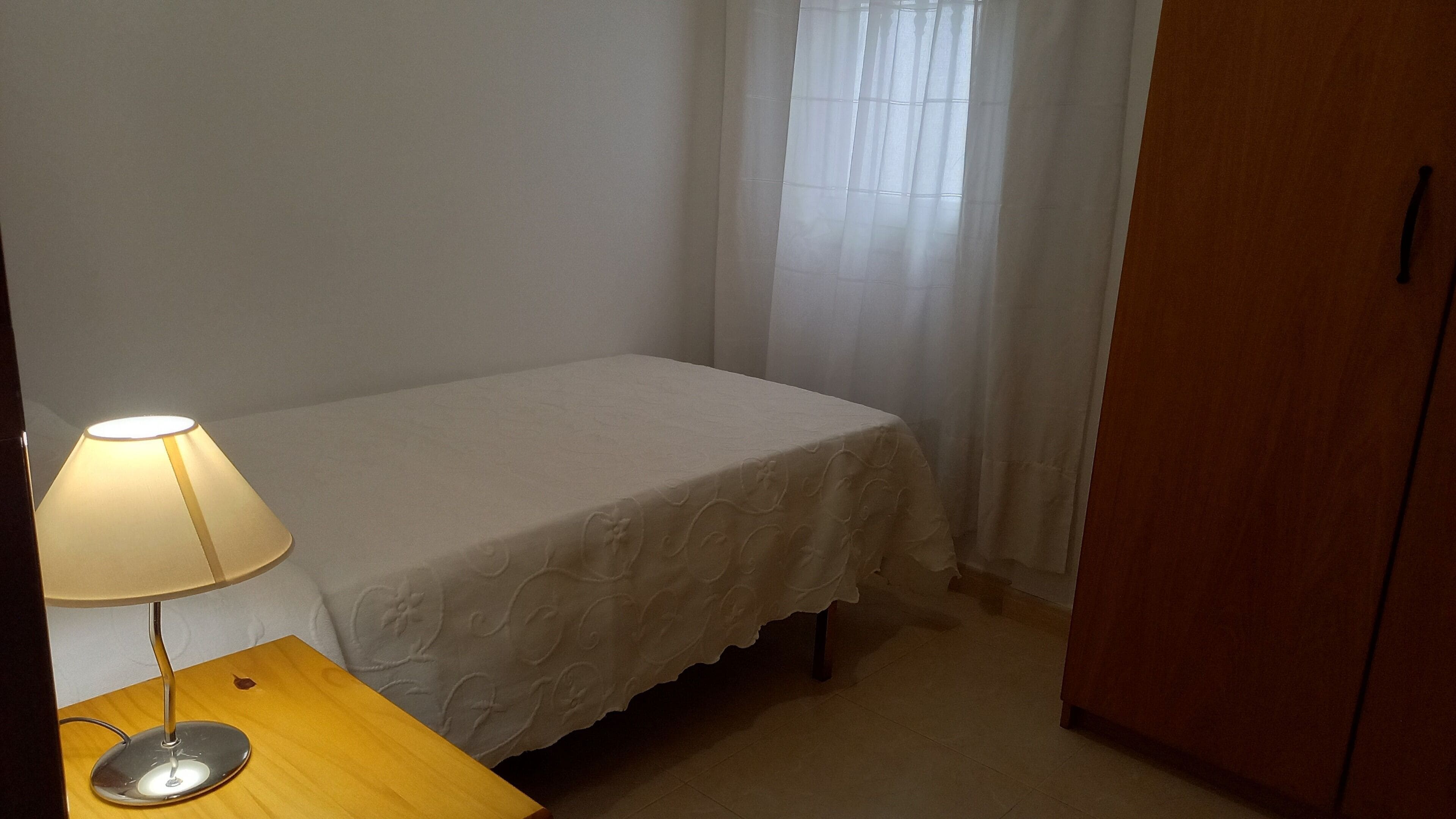 Una cama pequeña y bien hecha con una colcha blanca, una lámpara de noche y una mesita de madera.