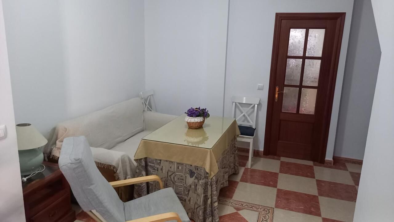 A small, compact living space with a sofa, a table with a patterned tablecloth, a chair, a lamp, and a door. - Andalusia Tourist Accommodation (Córdoba)
