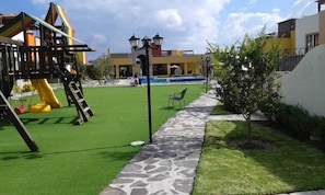 A playground with a slide and climbing structure, a pool, and a pathway leading to a seating area.