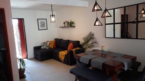 Un salon moderne avec un canapé d’angle, une table à manger avec des sets de table, des luminaires suspendus et une étagère murale agrémentée d’objets décoratifs.