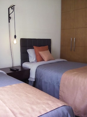 A bedroom with two beds, a nightstand, a wardrobe, and a wall-mounted light fixture.