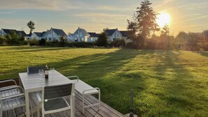 Eine Terrasse mit Tisch und Stühlen, mit Blick auf eine Wiese, im Hintergrund Häuser und ein untergehender Sonnenuntergang.