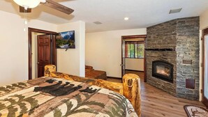 A bedroom with a large bed, a stone fireplace, a ceiling fan, and a television on the wall.