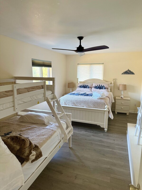 A bedroom with bunk beds, a ceiling fan, a window with blinds, a bedside table with a lamp, and a wall decoration.