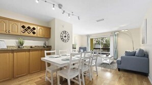 Une cuisine moderne avec des meubles en bois, un coin repas avec des chaises blanches et une table, et un salon avec un canapé et une grande fenêtre.