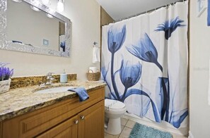 Et badeværelse med bordplade i granit, hvid vask, trækommode, hvid toilet og bruseforhæng med blomstermønster.