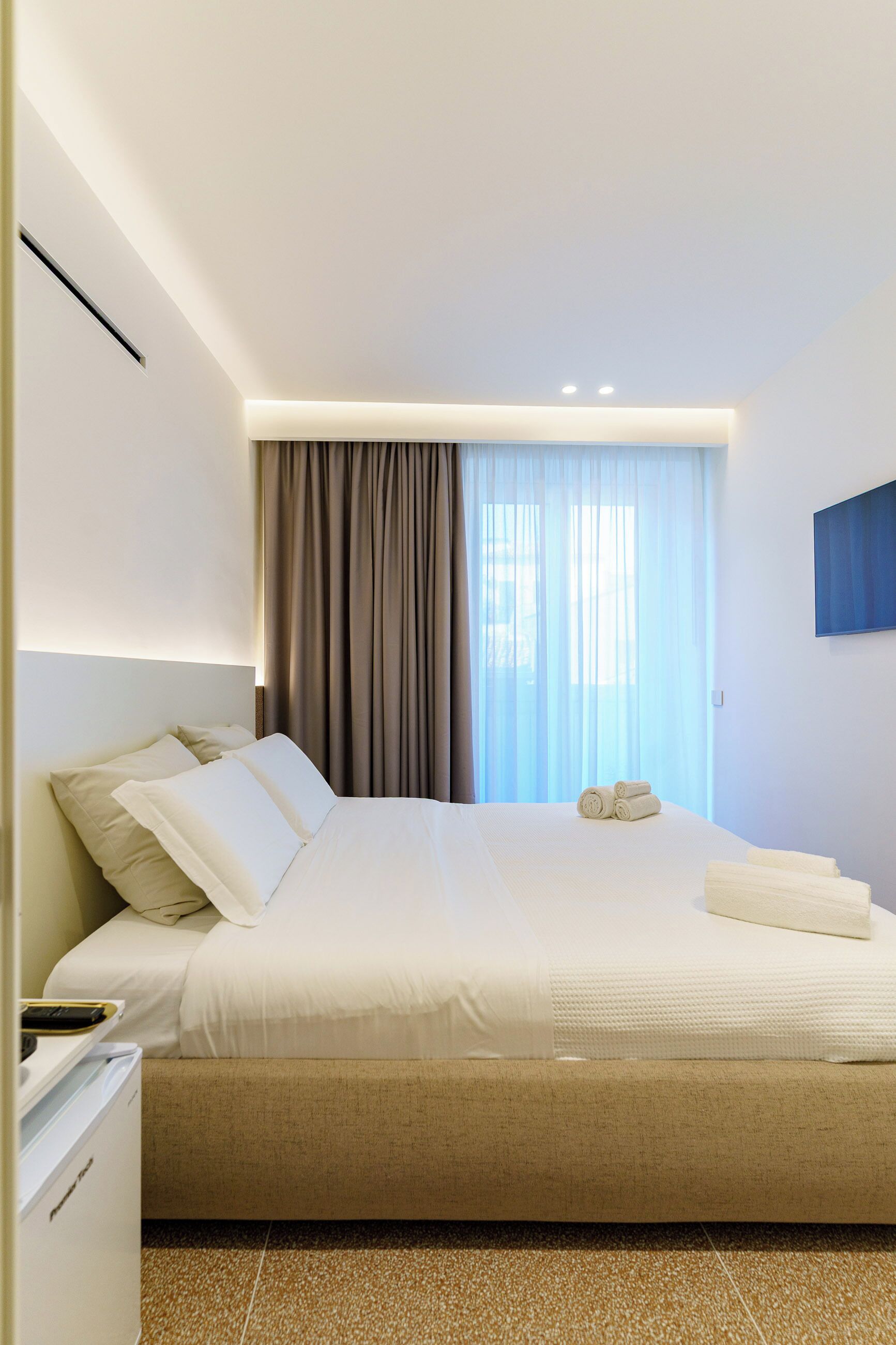 A modern hotel room with a large bed, a bedside table, a built-in desk, and a window with curtains.