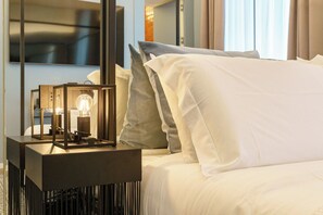 Ein ordentlich bezogenes Bett mit weißen und grauen Kissen, ein Nachttisch mit einer Lampe und ein moderner, an der Wand befestigter Fernseher.