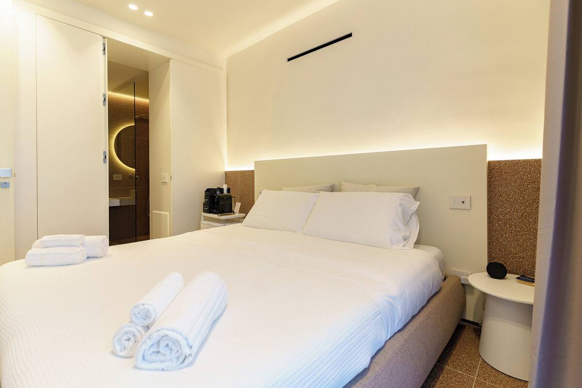 A hotel room with a neatly made bed, a bedside table with a lamp, and a wall-mounted light fixture.