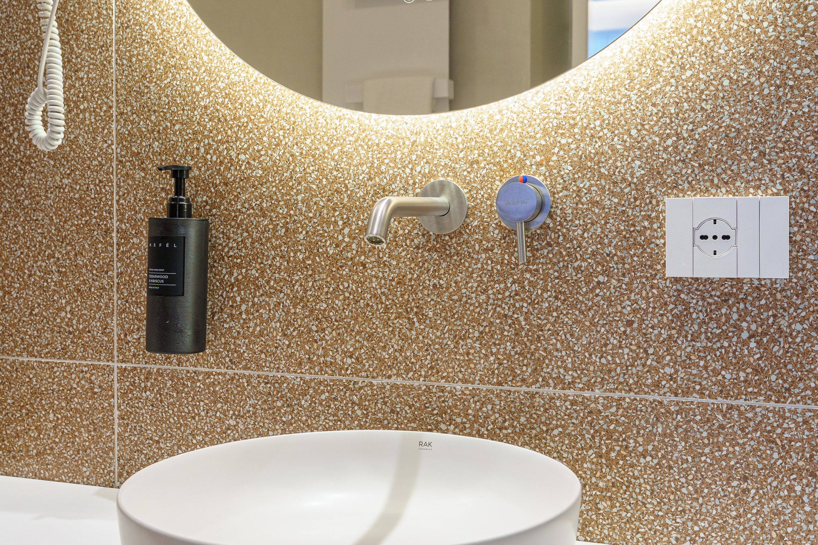 A bathroom with a speckled tile wall, a white sink, a black soap dispenser, a faucet, and a power outlet.