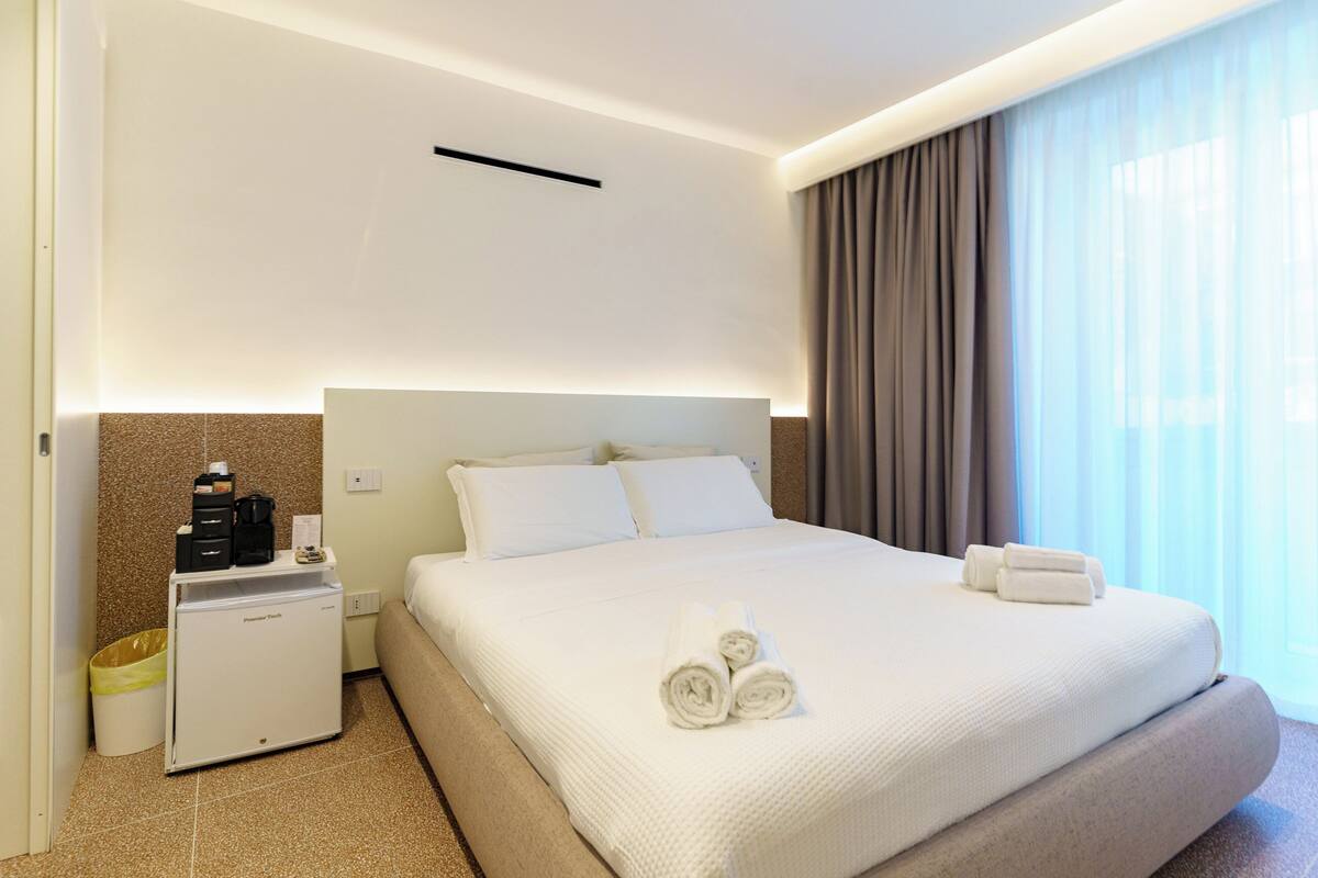 A hotel room with a bed, a mini-fridge, a trash bin, and a window with curtains.