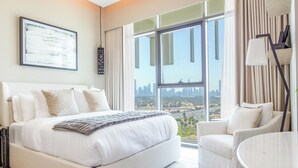A hotel room with a large bed, a sitting area with a chair and a table, a city view through large windows, and a framed artwork on the wall. - Bloomfields 1BR Azizi Farhad Burj View (Dubai)