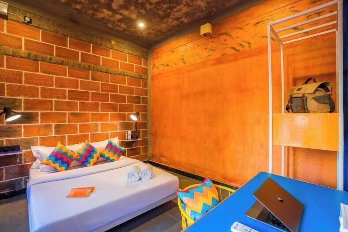 A room with a brick wall, a bed with colorful pillows, a blue table with a laptop, and a shelf with a bag.