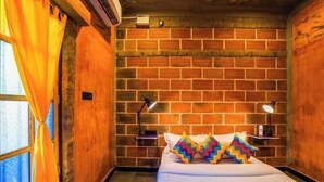 A hotel room with a brick wall, a bed with colorful pillows, a nightstand with a lamp, and a window with curtains. - goSTOPS Pondicherry Near Auroville (Auroville)