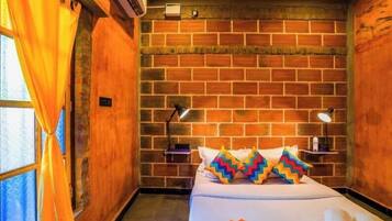 Quarto de hotel com parede de tijolos, cama com almofadas coloridas, mesa de cabeceira com abajur e janela com cortinas.