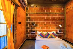 A hotel room with a brick wall, a bed with colorful pillows, a nightstand with a lamp, and a window with curtains. - goSTOPS Pondicherry Near Auroville (Auroville)