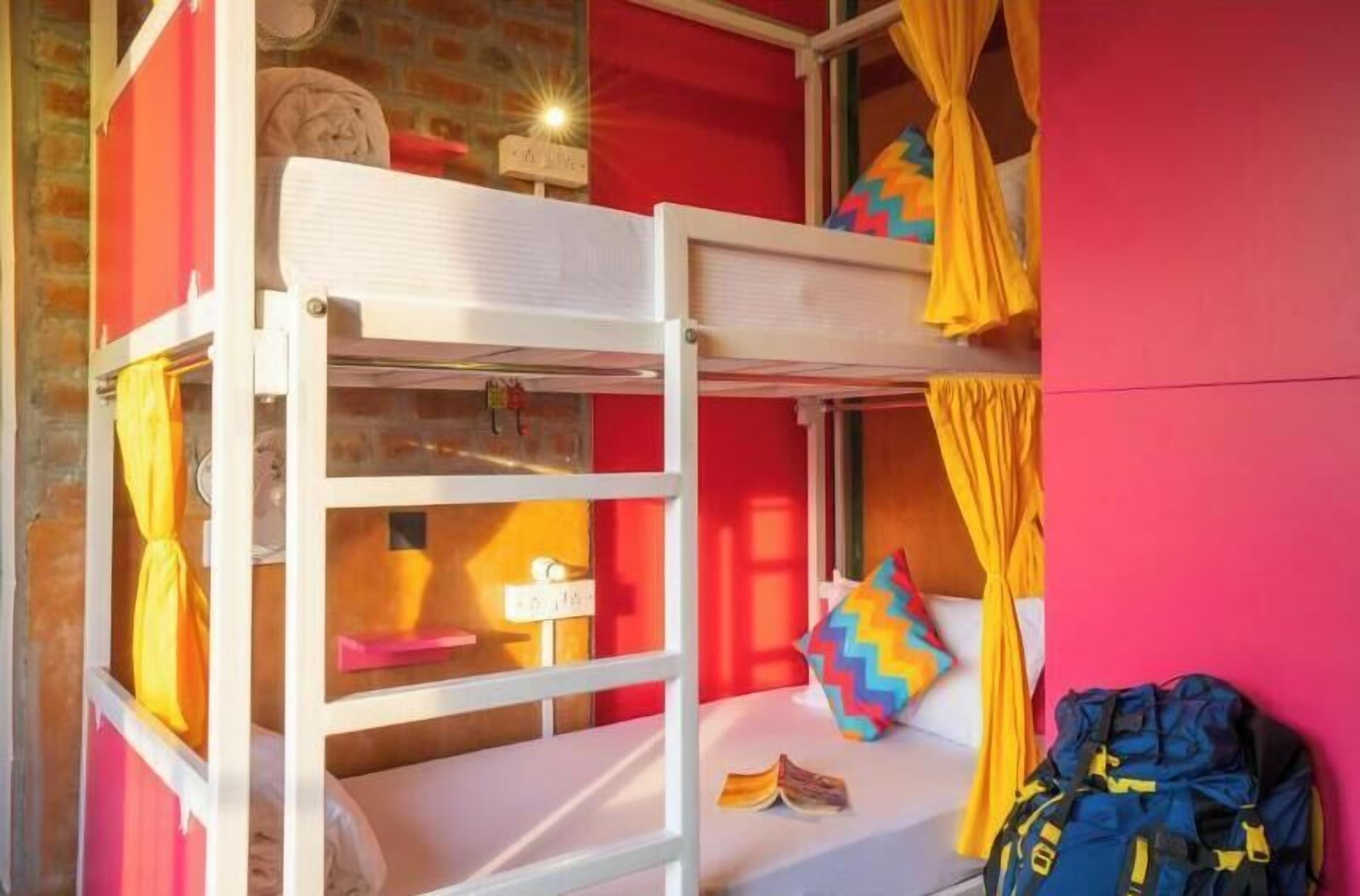 Een slaapkamer met een stapelbed, een bakstenen muur, een raam met gordijnen en een rugzak op de vloer.