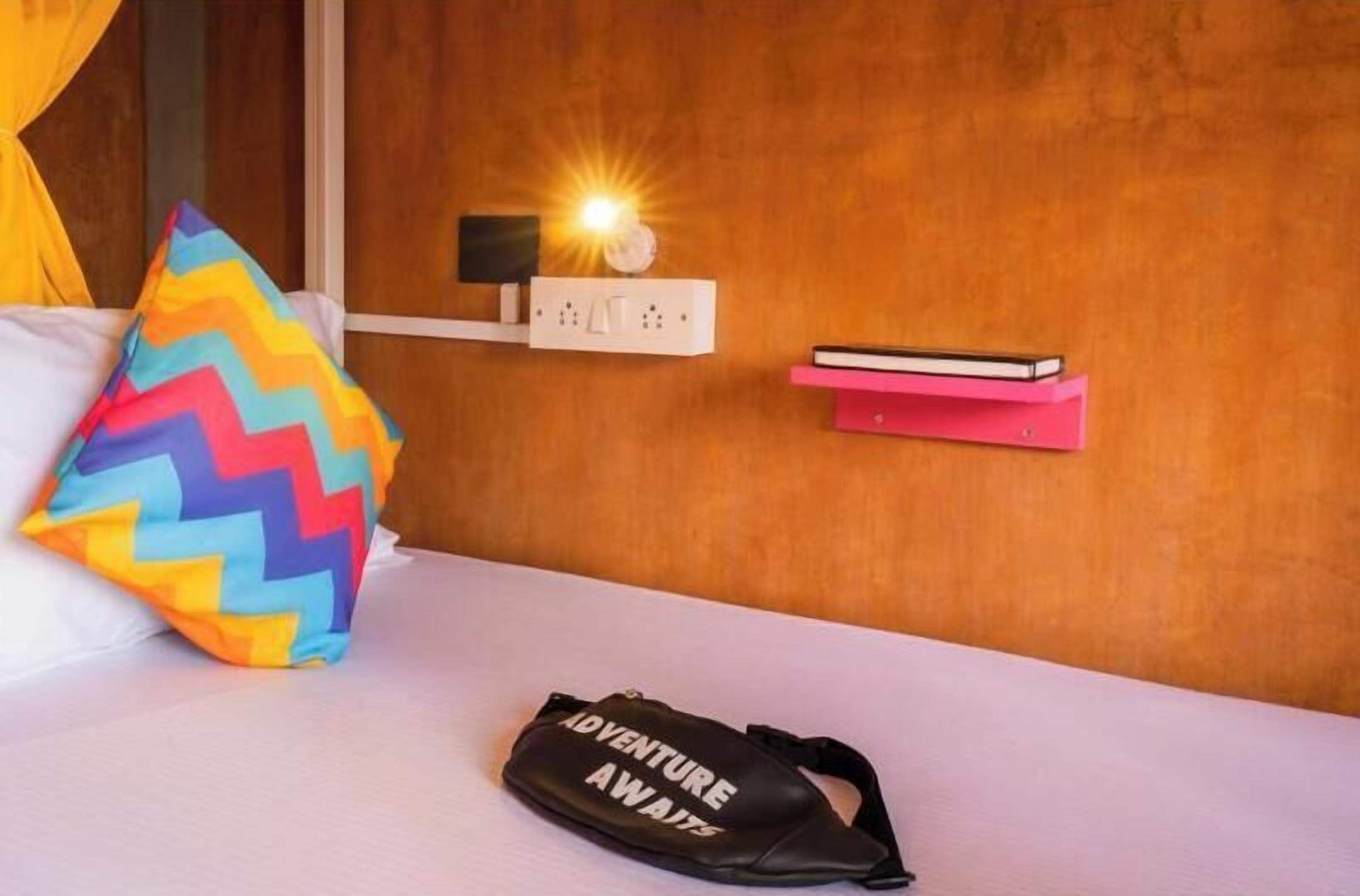 Un letto con un cuscino colorato a zig-zag, una borsa nera con scritte e un comodino con un libro e una lampada.