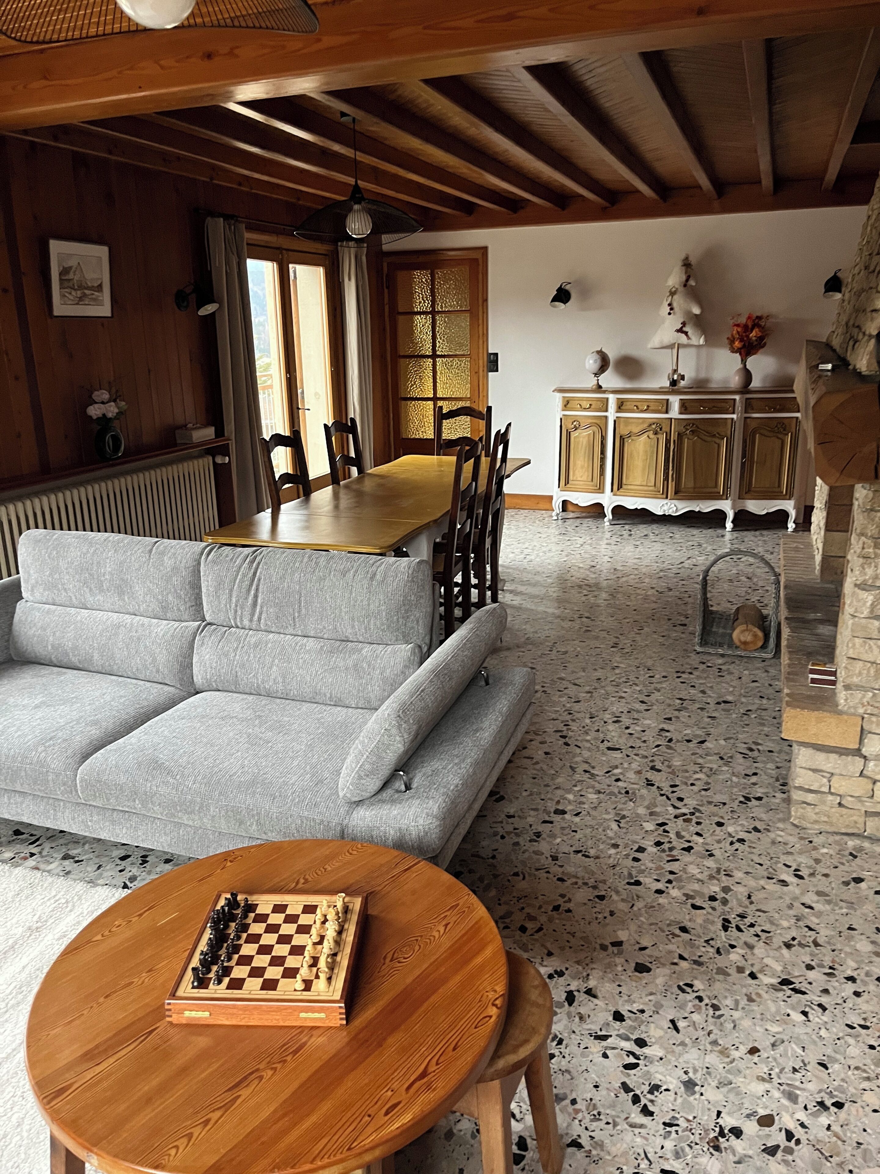 A cozy living room with a grey sofa, wooden dining table, and a chessboard on a round wooden table.