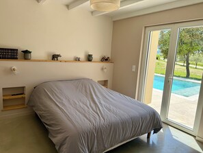 A bedroom with a bed, a wooden shelf with decorative items, a small window, and a view of a swimming pool. - Large, renovated Provençal farmhouse with a pool (Valréas)
