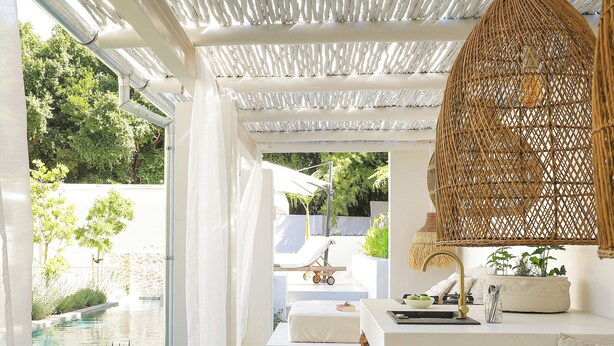 A modern outdoor seating area with a wooden table and chairs, a wicker hanging light fixture, and a view of a pool and greenery. - Bel Ombre Hills, 2200m2 luxury property in upper Constantia (Cape Town)