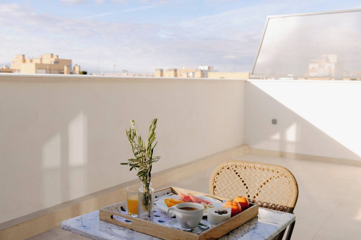 A breakfast tray on a balcony with a wicker chair, a vase with greenery, and a cityscape in the background.