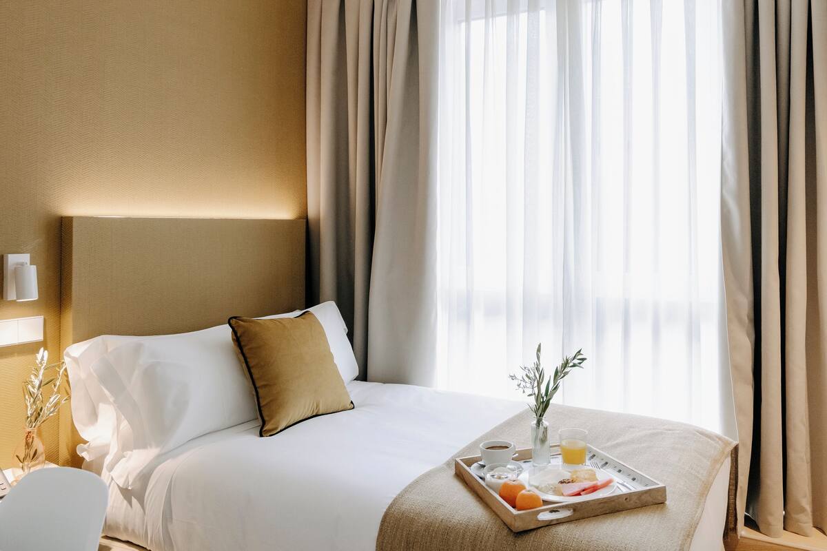 A hotel room with a neatly made bed, a tray with breakfast items, and a window with sheer curtains.