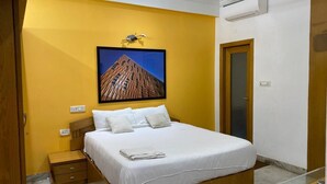 A hotel room with a bed, a nightstand, a wooden door, and a wall-mounted air conditioner. - Mithra Homes (Hyderabad)