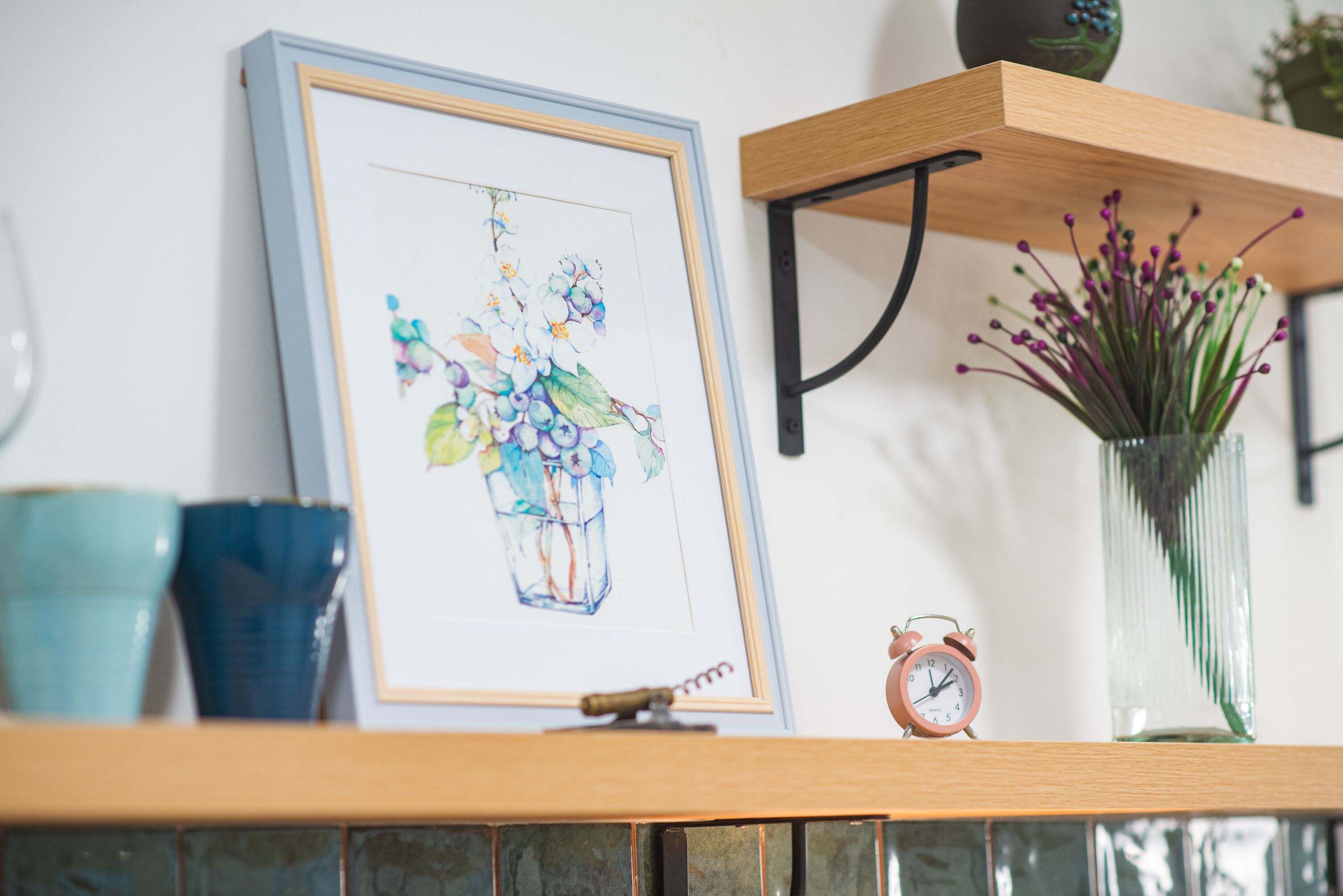 A framed artwork of flowers on a shelf, a clock, and a vase with purple flowers.