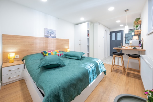 A modern bedroom with a wooden headboard, a bed with a teal bedspread, a bedside table with a lamp, and a small desk with stools. - HB apartments Irakli Abashidze (Tbilisi)
