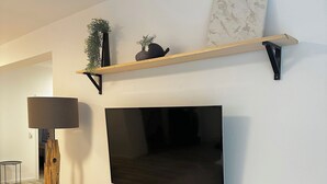 Un salon moderne avec un téléviseur à écran plat fixé au mur, une table basse en bois avec une télécommande, un lampadaire et une étagère ornée d’objets décoratifs.