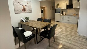 Un coin repas avec une table en bois et des chaises noires, une cuisine avec des meubles blancs et un téléviseur, et un tableau représentant un cerf accroché au mur.