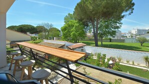 A balcony with a wooden table and chairs, a view of a green lawn, and a large tree. - Les Salines holiday home 5 minutes from the beach (Le Grau-du-Roi)