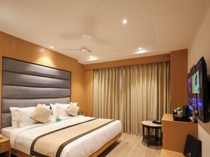 A hotel room with a large bed, a bedside table, a desk with a chair, a television, and a ceiling fan.