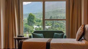 A hotel room with a bed, a chair, a small table, and a large window offering a view of a mountainous landscape. - Click Hotel Bhuntar,Kullu-Bhuntar Airport (Aut)