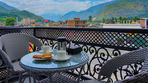 A balcony with wicker chairs and a glass table, overlooking a town with mountains in the background. - Click Hotel Bhuntar,Kullu-Bhuntar Airport (Aut)