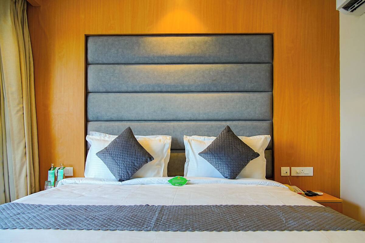 A hotel room with a large bed, two pillows, a bedside table with a remote control, and a nightstand with a bottle.