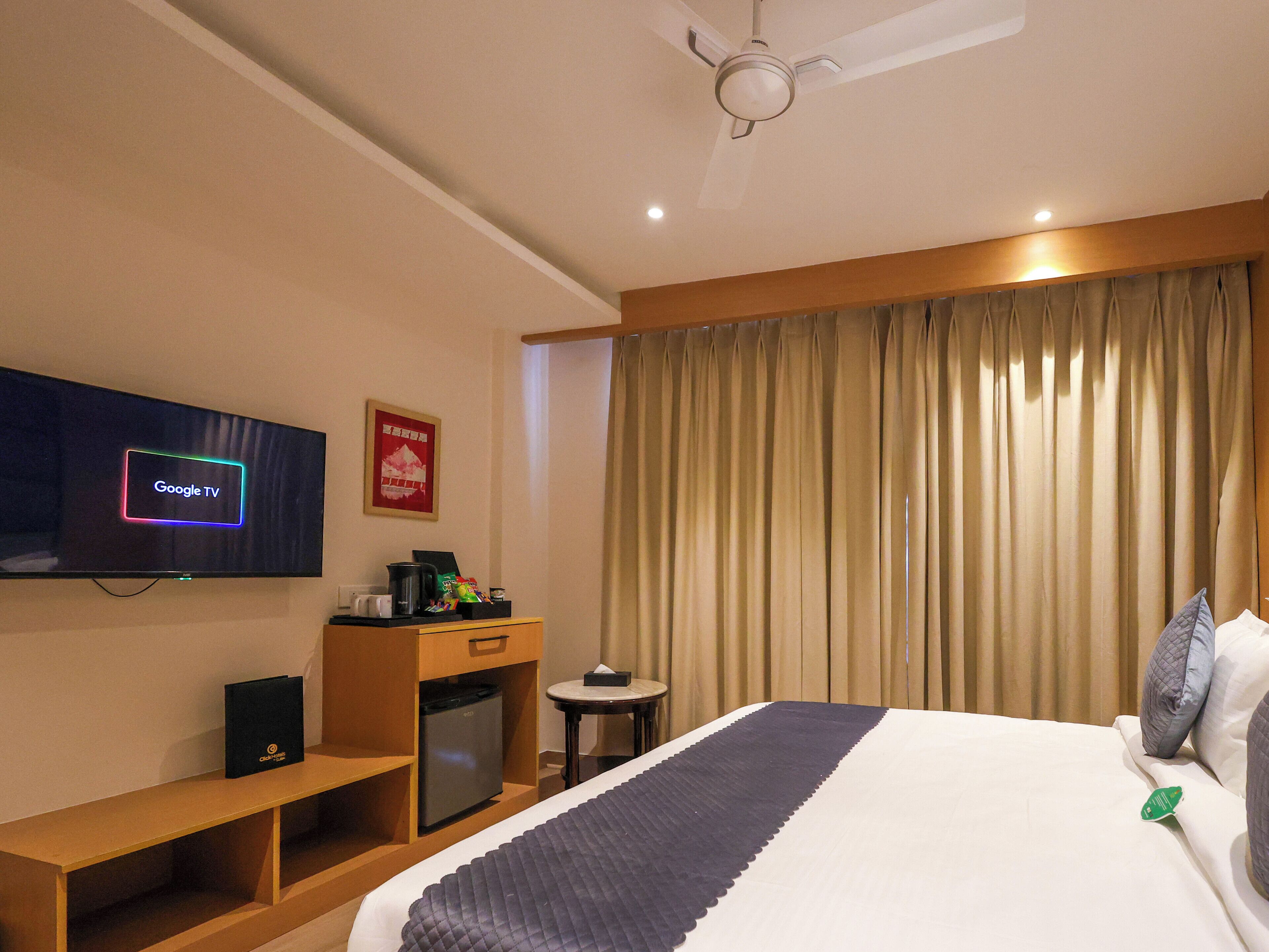 Habitación de hotel con cama, televisor mostrando 'Google TV', una mesita con tetera y cafetera, un refrigerador pequeño y una cortina.
