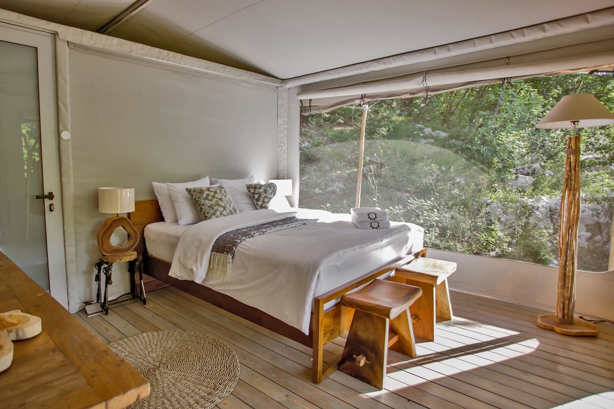 Une chambre à coucher avec un lit, une table de chevet, une lampe et une vue sur la verdure à l’extérieur.