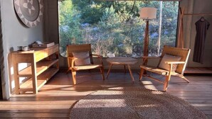 A cozy room with a wooden floor, a round table, two wicker chairs, a hanging lamp, and a view of a forest through large windows. - Ashuma retreat center (Cetinje)
