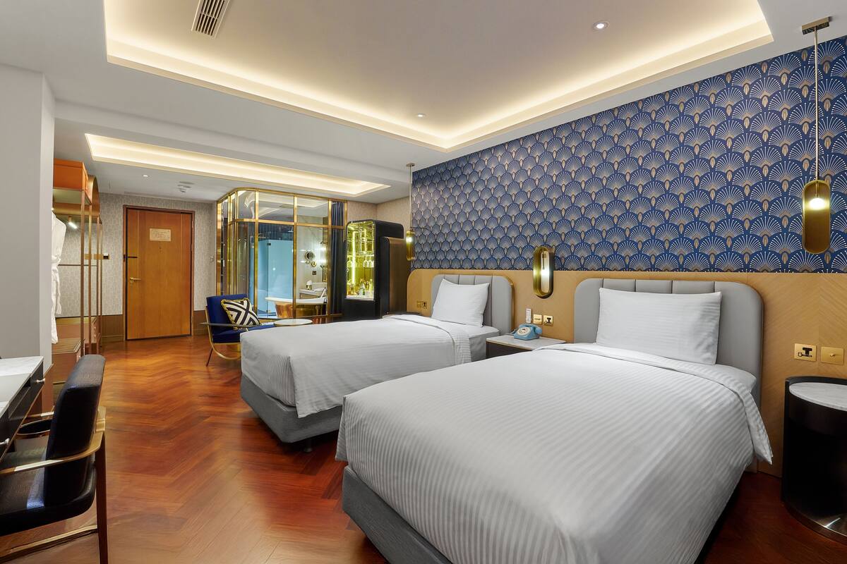 A hotel room with two beds, a desk, a chair, a mirror, and a patterned wall.