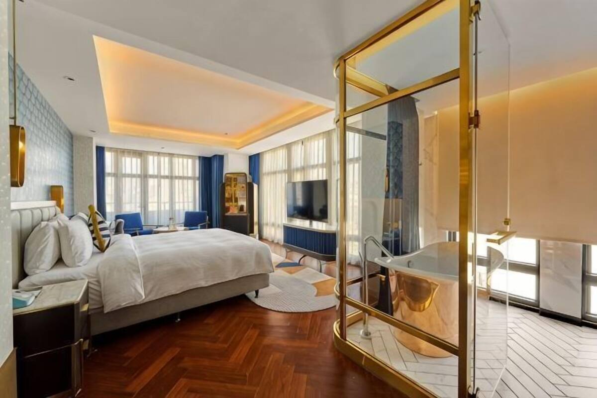 A modern hotel room with a large bed, a glass partition, a TV, and a view of the city.