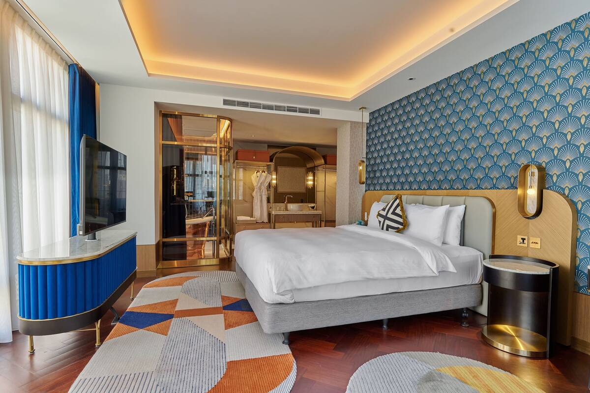 A modern hotel room with a large bed, a TV, a bathroom with a mirror, and a patterned wall.