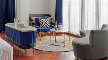 A modern hotel room with a marble bar, a grey armchair, a blue sofa with a black and white pillow, and a round table with a wine bottle and glasses.