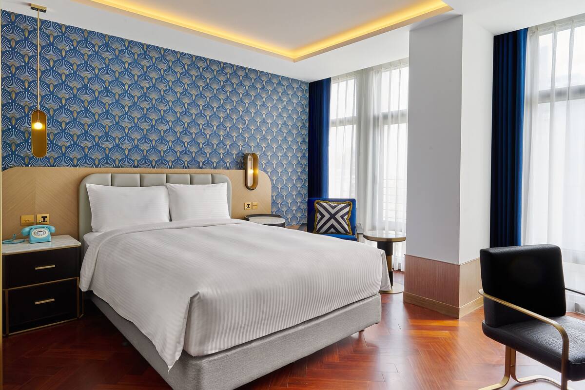 A hotel room with a large bed, bedside tables, a nightstand, a chair, a small table, and a patterned wallpaper.