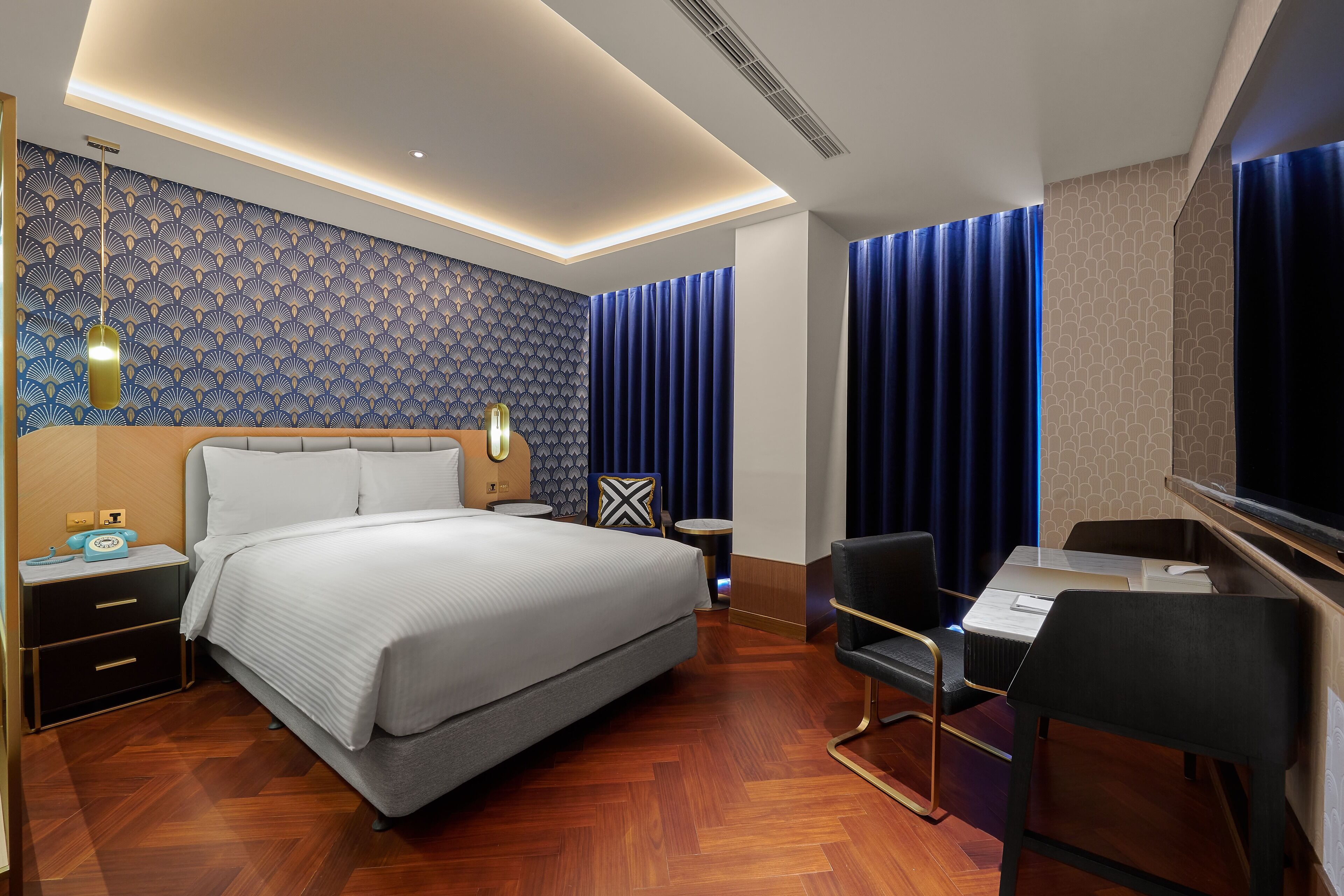 A modern hotel room with a large bed, a desk with a chair, a nightstand, and a wall with a geometric pattern.