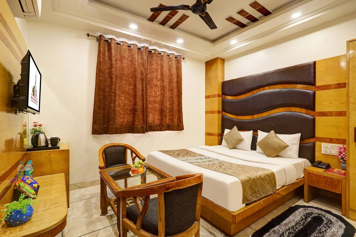 A hotel room with a bed, a desk with a chair, a small table with fruit, a ceiling fan, and a wall-mounted TV.