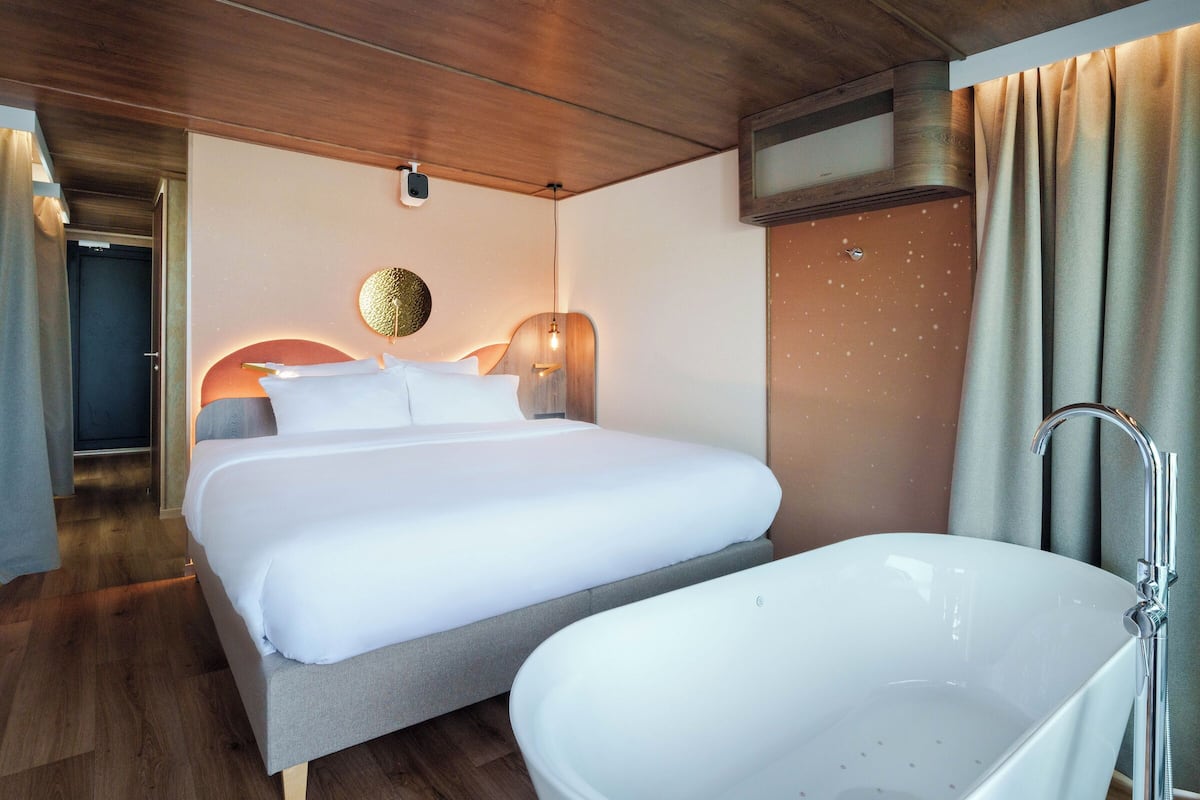 A modern hotel room with a large bed, a bathtub, and a wooden ceiling.