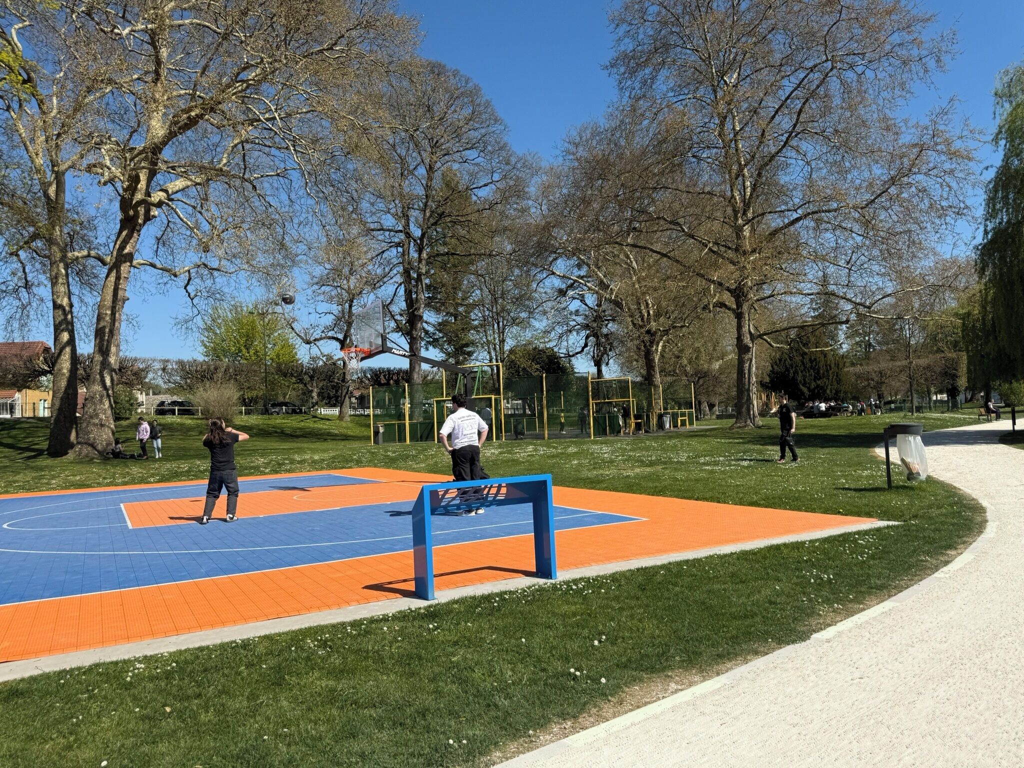 Una cancha de baloncesto con superficie azul y naranja, rodeada de árboles y un cielo despejado.