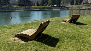 Dos sillones de madera junto a un lago tranquilo, bajo un cielo despejado, rodeado de vegetación y edificios residenciales.